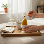 Bienenprodukte Shop Bienenwachskerze in Eiform, die von Hand mit einem Streichholz auf einem Holztablett angezündet wird.